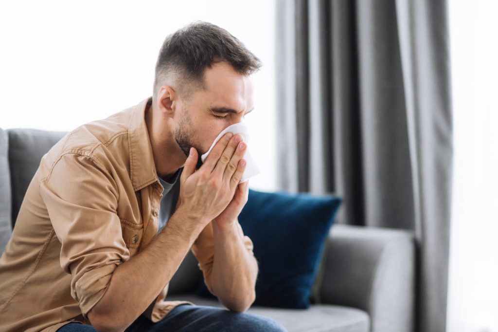 Veteran suffering from allergic rhinitis sitting on his couch blowing his nose.